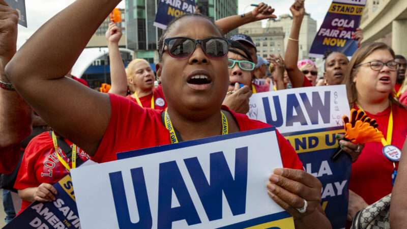 A woman wearing sunglasses holds up a UAW protest sign with other protesters who are also wearing red shirts.