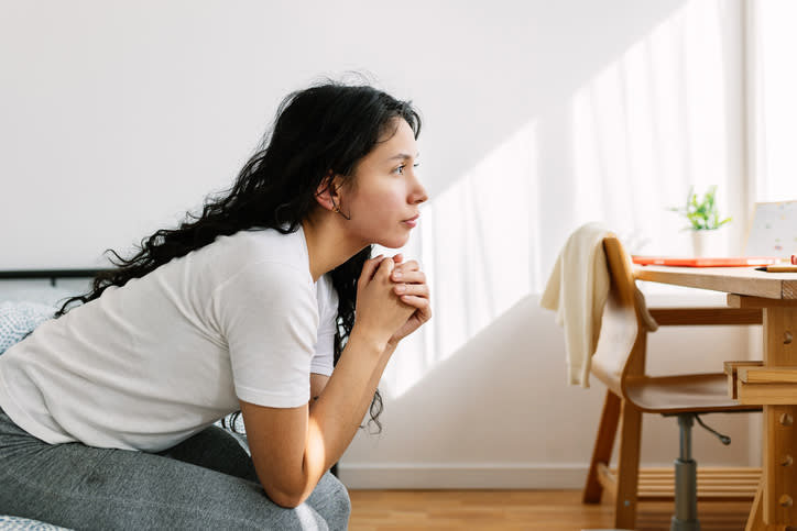 Woman in casual attire rests chin on hands, looking contemplatively out a window from a cozy indoor setting