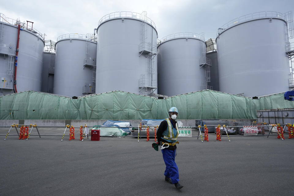 A worker helps direct a truck driver as he stands near tanks, background, that will be used to store treated radioactive water after it was used to cool down melted fuel at the Fukushima Daiichi nuclear power plant, run by Tokyo Electric Power Company Holdings (TEPCO), in Okuma town, northeastern Japan, Thursday, March 3, 2022. Treated radioactive water is stored in tanks at the power station. The government has announced plans to release the water after treatment and dilution to well below the legally releasable levels through a planned undersea tunnel at a site about 1 kilometer offshore. (AP Photo/Hiro Komae)