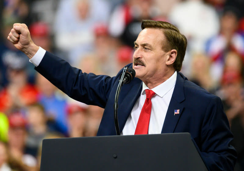 A mustached man in a dark suit, white shirt and red tie holding his right fist up in front of a crowd