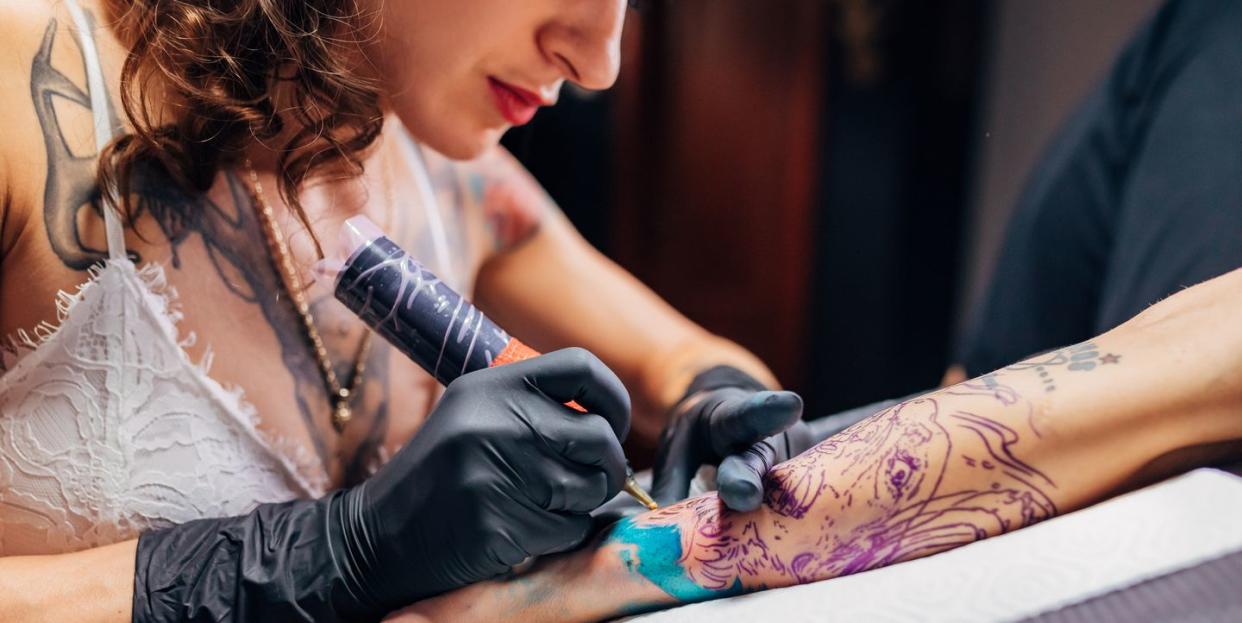 tattoo artist wearing black latex gloves tattooing colourful tattoo on woman's visible outstretched arm