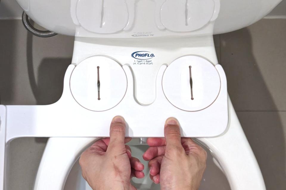 A person using both hands to lower a Tushy Classic bidet onto a two-piece toilet bowl with the seat removed