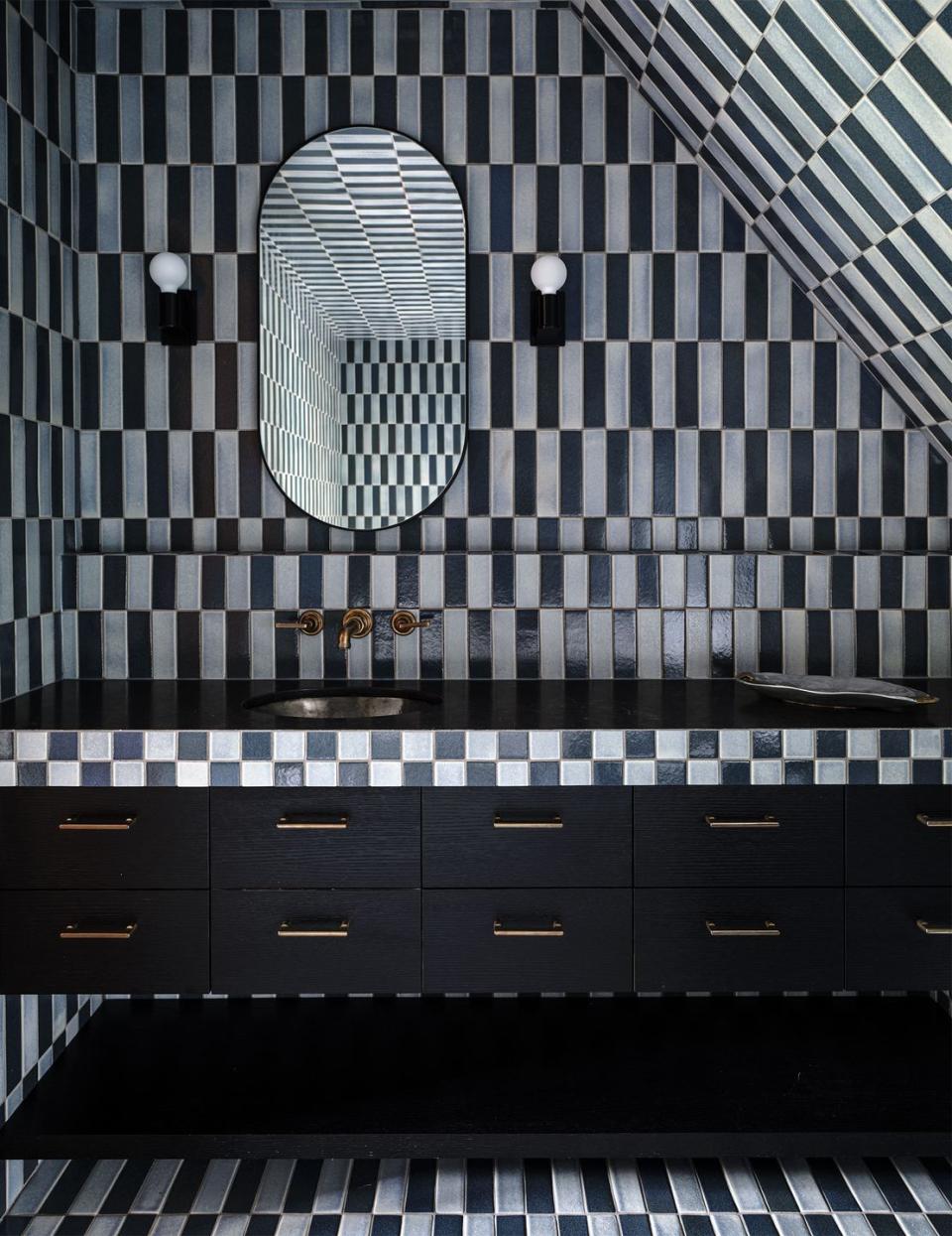 black and white rectangular tiles cover bathroom walls, floor, counter with sink and drawers, long oval mirror, two small globe sconces flank the mirror