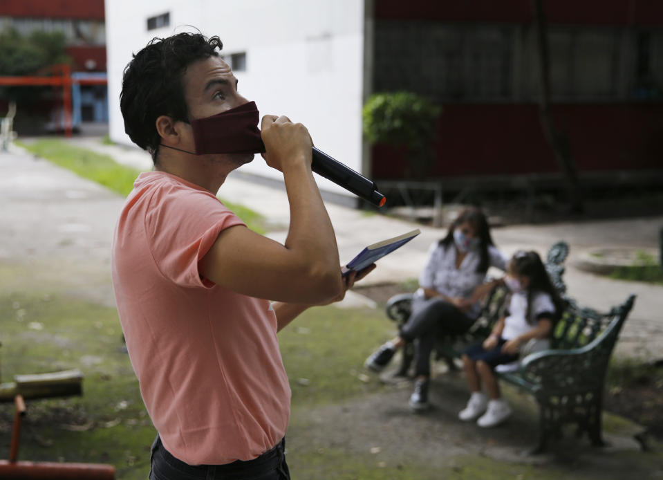 Residents listen from a bench, background, as Percibald Garcia reads children's books aloud outside the high-rise buildings in the Tlatelolco housing complex, in Mexico City, Saturday, July 18, 2020. Confinement during the coronavirus pandemic has been especially tedious for children, so the young architect sets out every afternoon with a microphone and a loudspeaker to walk the neighborhood where he lives and provide entertainment. (AP Photo/Marco Ugarte)