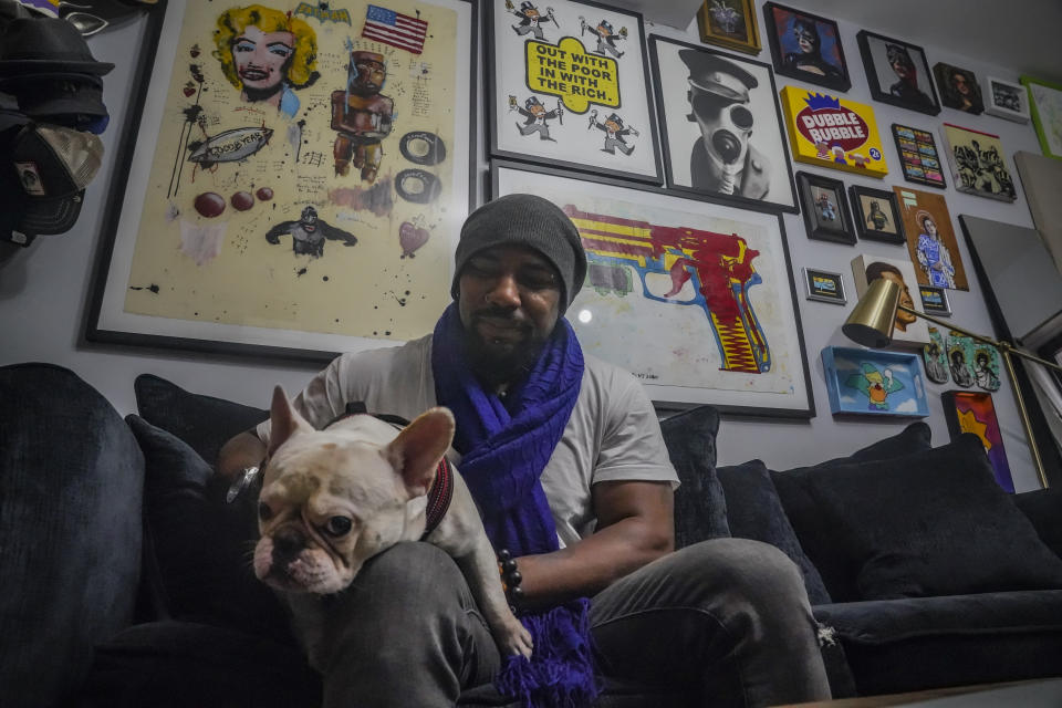Painter Guy Stanley Philoche, a 43-year-old Haitian immigrant and star in the New York art world, sits with his dog Picasso at their East Harlem home next to a wall of art collected from other artist, Thursday Nov. 19, 2020, in New York. After a hugely successful gallery show, Philoche wanted to treat himself to a fancy $15,000 watch, instead he bought the works of fellow artists struggling in the pandemic. "I'm not a rich man," he said, "but I owe a big debt to the art world." (AP Photo/Bebeto Matthews)