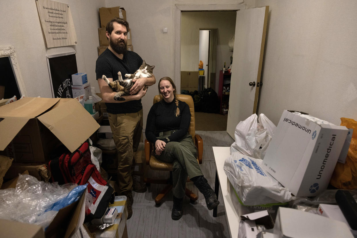 Crow cradles a cat and Rigan sits in an office chair in a room with boxes of supplies.