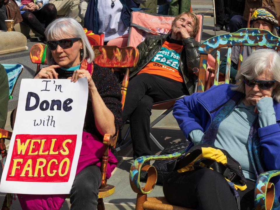 older women sit in rocking chairs in the sun holding sign reading i'm done with wells fargo
