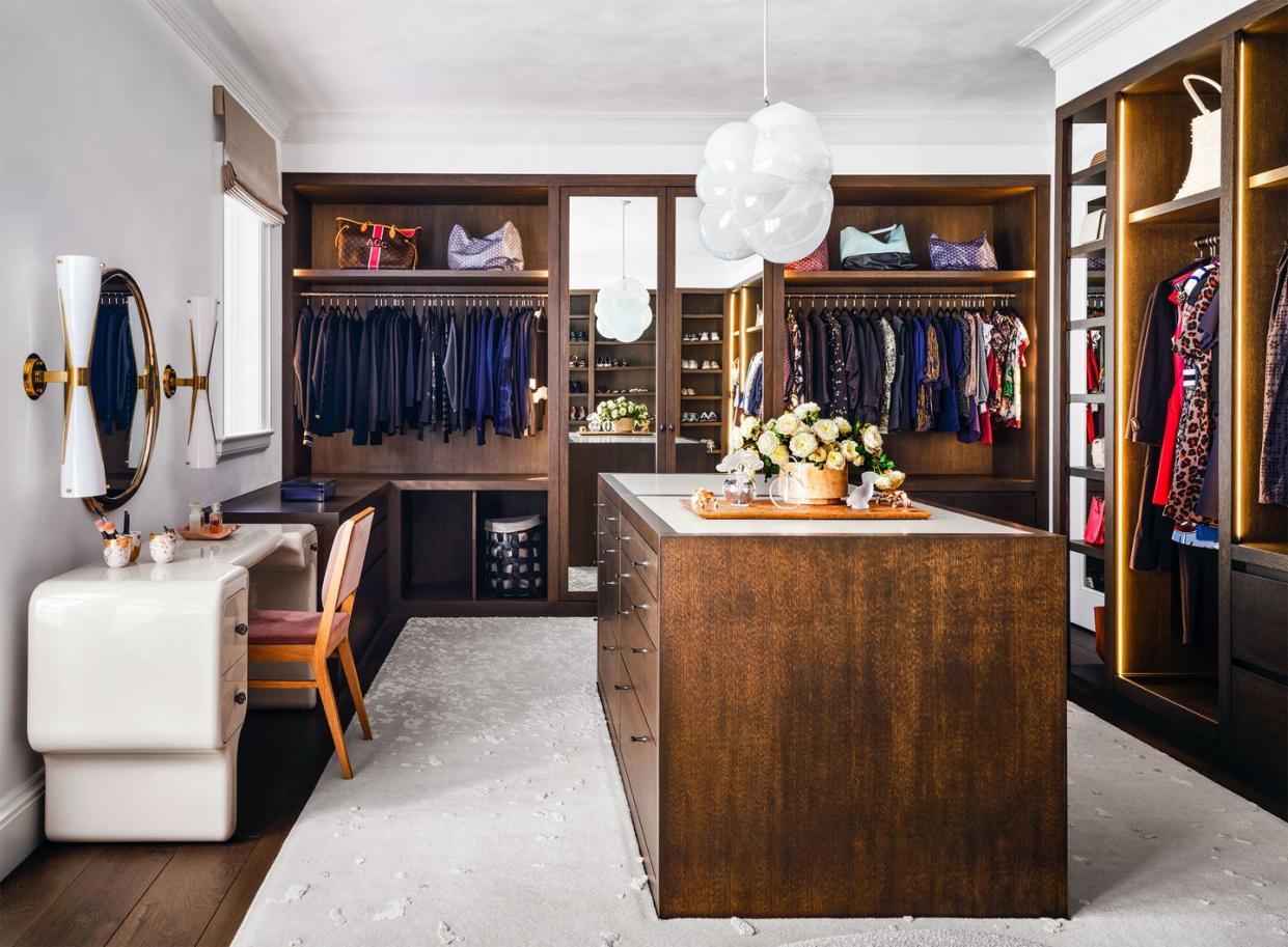 in the dressing room at left sits a vanity with a mirror and sconces, in the center is a large chest of drawers, and in the back and along right are open closets for hanging clothing and compartments for storage