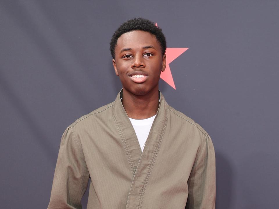 alex hibbert, a young man with shirt natural hair, in off-white linen pants, a loose fitting green shirt, smiling on a red carpet