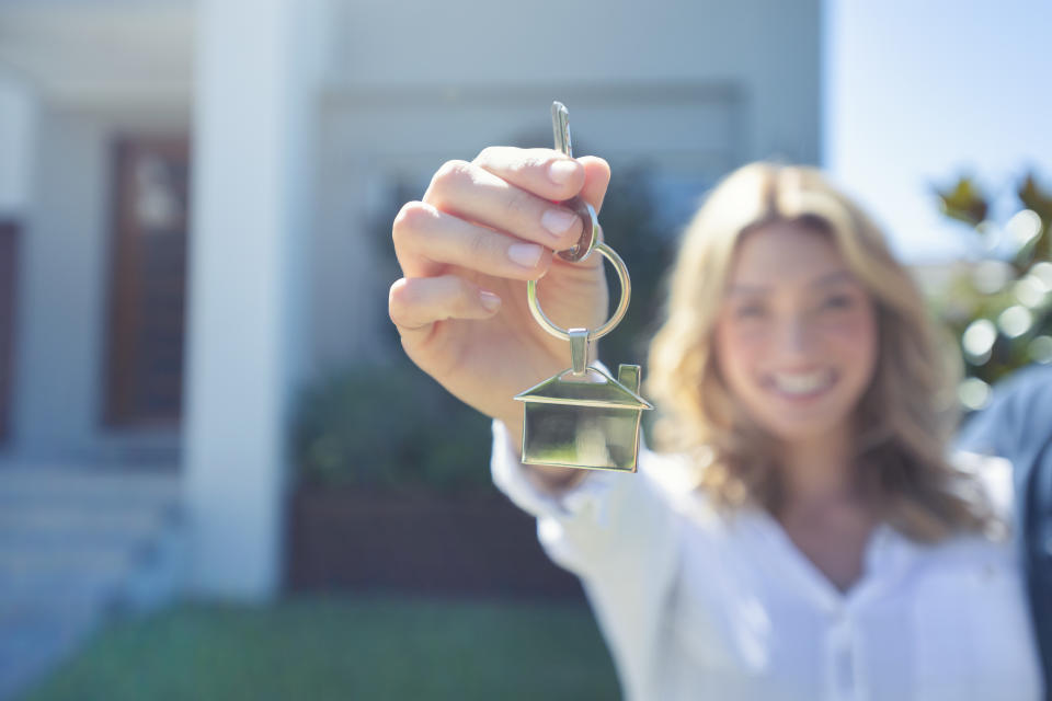 Young woman holding the key to her new house. She is smiling and happy. They keyring is shaped like a house. The house can be seen in the background. For a story about the First Home Savings Account.