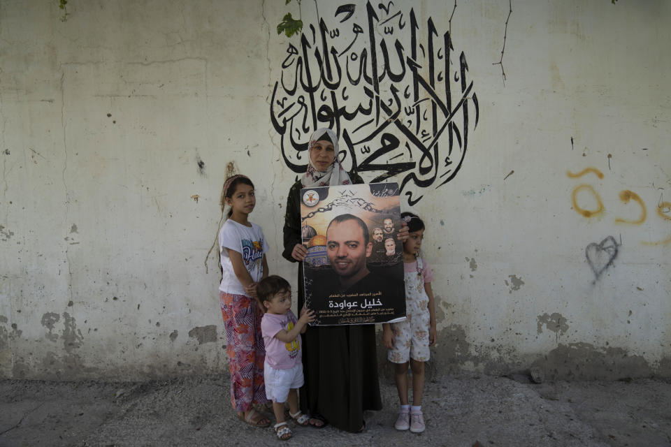 Family members of Khalil Awawdeh, a Palestinian prisoner in Israel, from right, Tolin, 9, Maryam, 21 months, his wife Dalal, 32, and Maria, 4 pose with a poster of Khalil Awawdeh at the family house, in the West Bank village of Idna, Hebron, Tuesday, Aug. 9, 2022. Arabic in the background reads "There is no God but Allah, Muhammad is the Messenger of Allah." Awawdeh who has refused food for 150 days and is wasting away in an Israeli jailhouse infirmary has suddenly been thrust at the center of efforts to firm up a Gaza cease-fire. (AP Photo/Nasser Nasser)