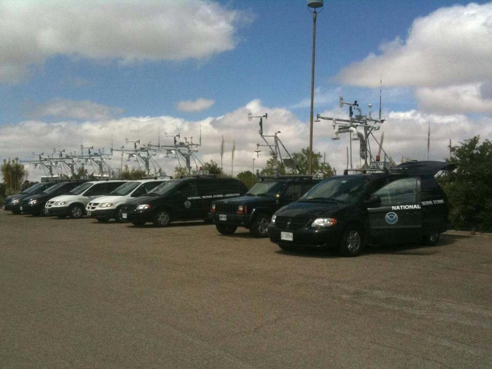 A row of seven minivans, SUVS and jeeps with racks on top holding the sorts of instruments one might see in a weather station.