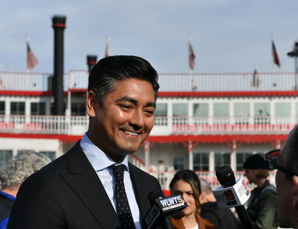 cincinnati major aftab pureval smiles as unseen people hold microphones toward him, he wears a suit and tie and stands outside