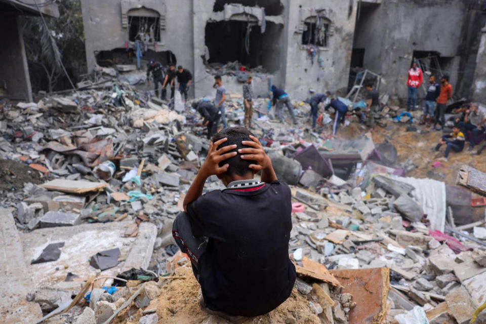 A person sits foreground, hands on head, facing the rubble of a collapsed building with onlookers amidst debris