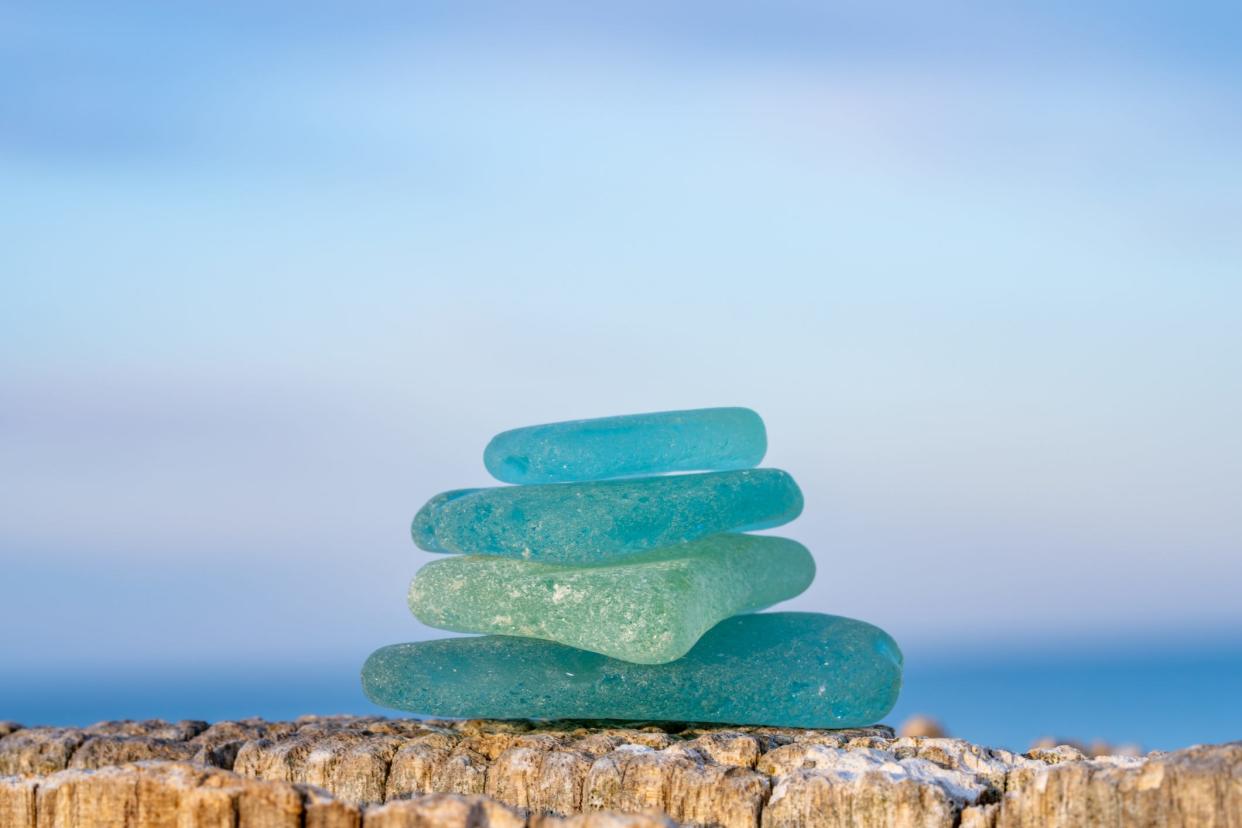 A pile of blue and green sea glass sits on a piece of driftwood on the beach.