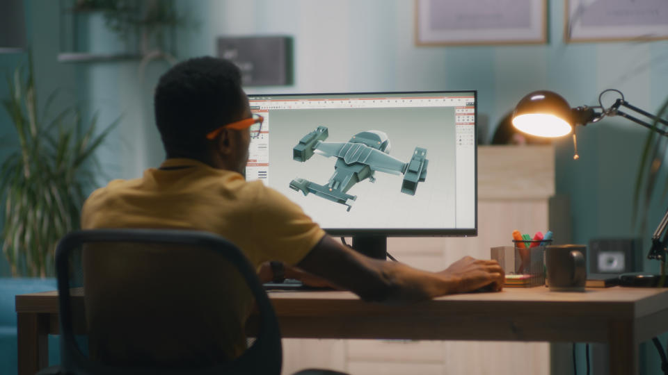 Back view of black man in yellow t shirt and glasses creating 3D model of modern aircraft on computer while sitting at table in home office and working on remote project