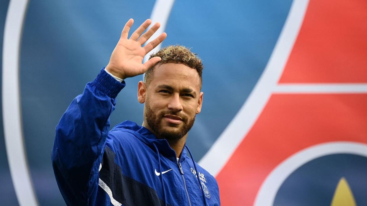 neymar waves and looks at the camera with a solemn expression, he wears a blue team jacket for paris saint germain