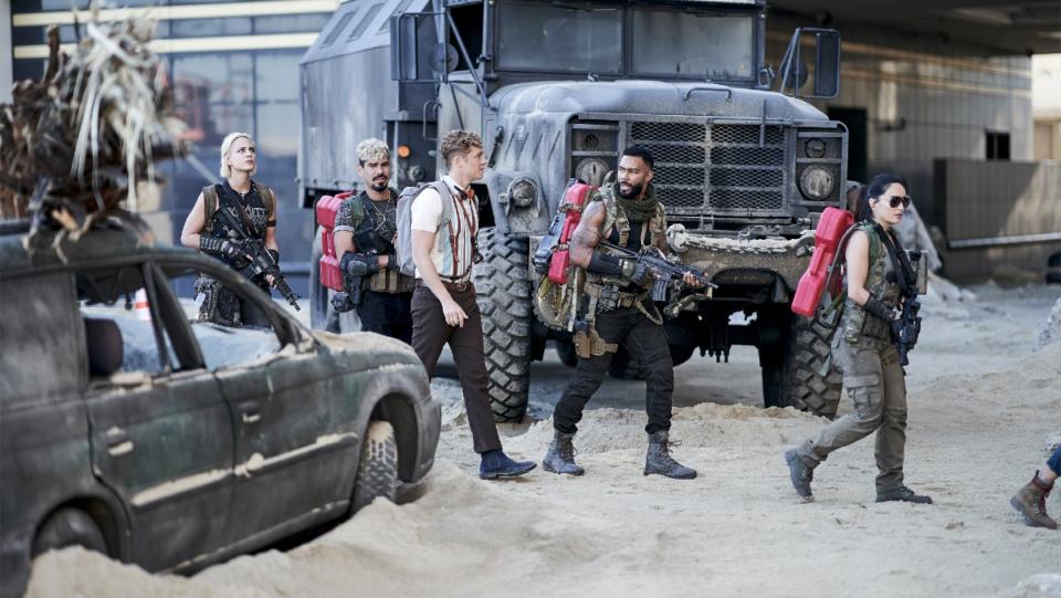 A heist team walks across sand holding weapons surrounded by abandoned cars in a still from Army of the Dead.