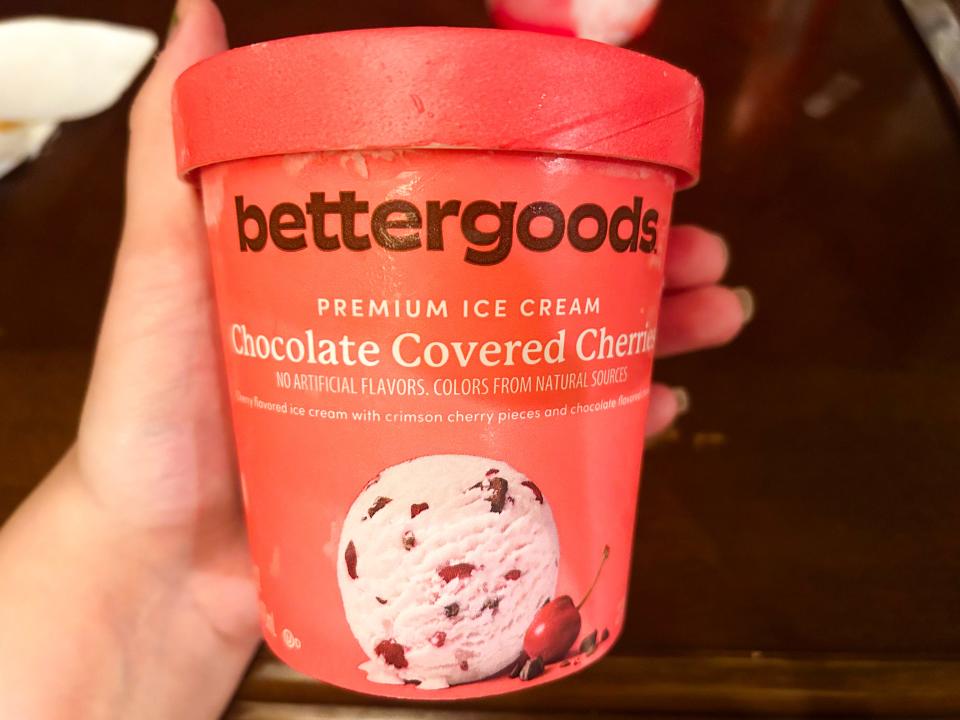 A hand holds a red-orange carton of Bettergoods chocolate-covered cherries ice cream with a scoop of ice cream image on the container