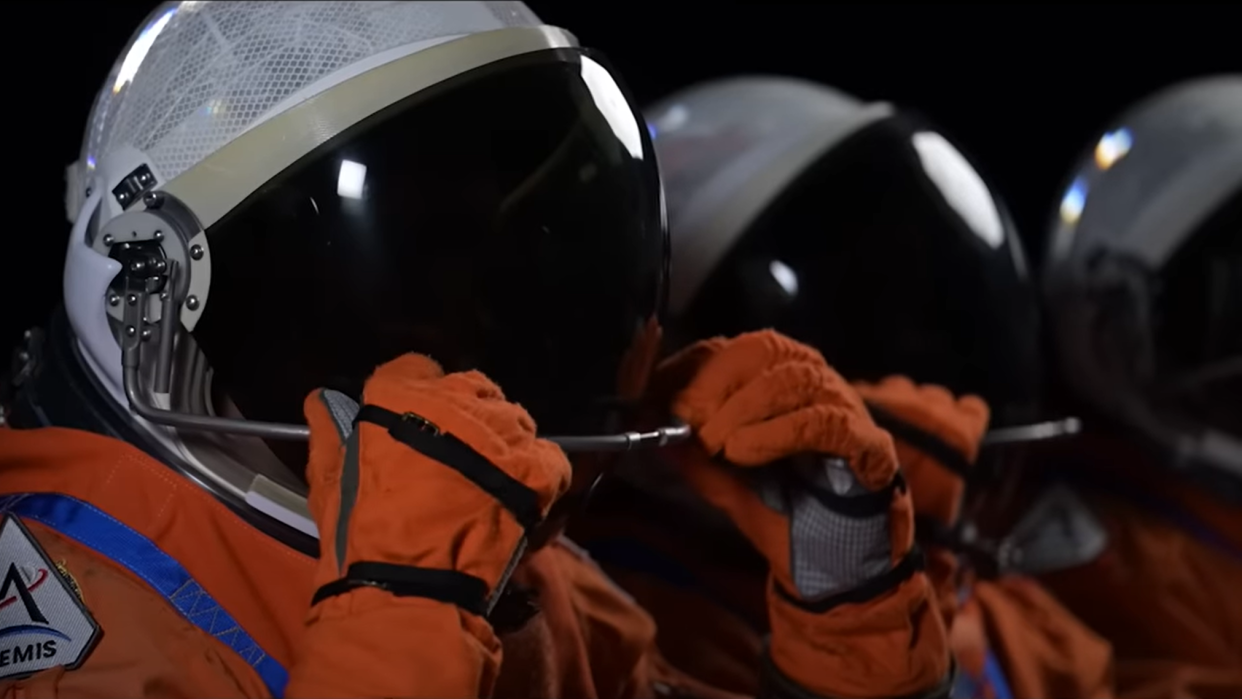  three astronauts in spacesuits with white helmets. their visors are black and we cannot see their face. the astronauts grasp their visors in a gesture that suggests their faces will be visible soon, once the visor is lifted 