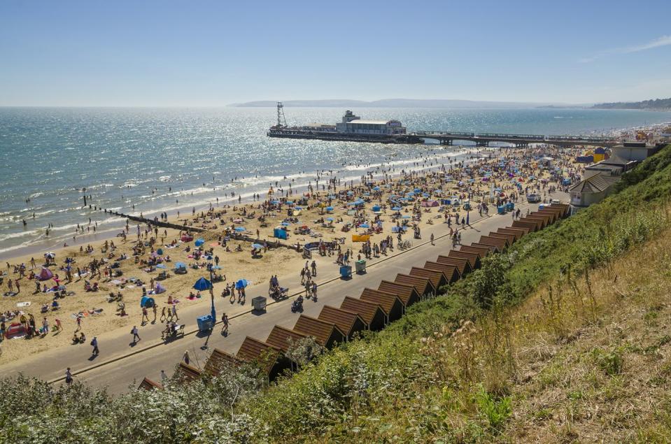 crowds of sunseekers on the sandy beach at bournemouth, with the pier in the distance, and beach huts in the foregound