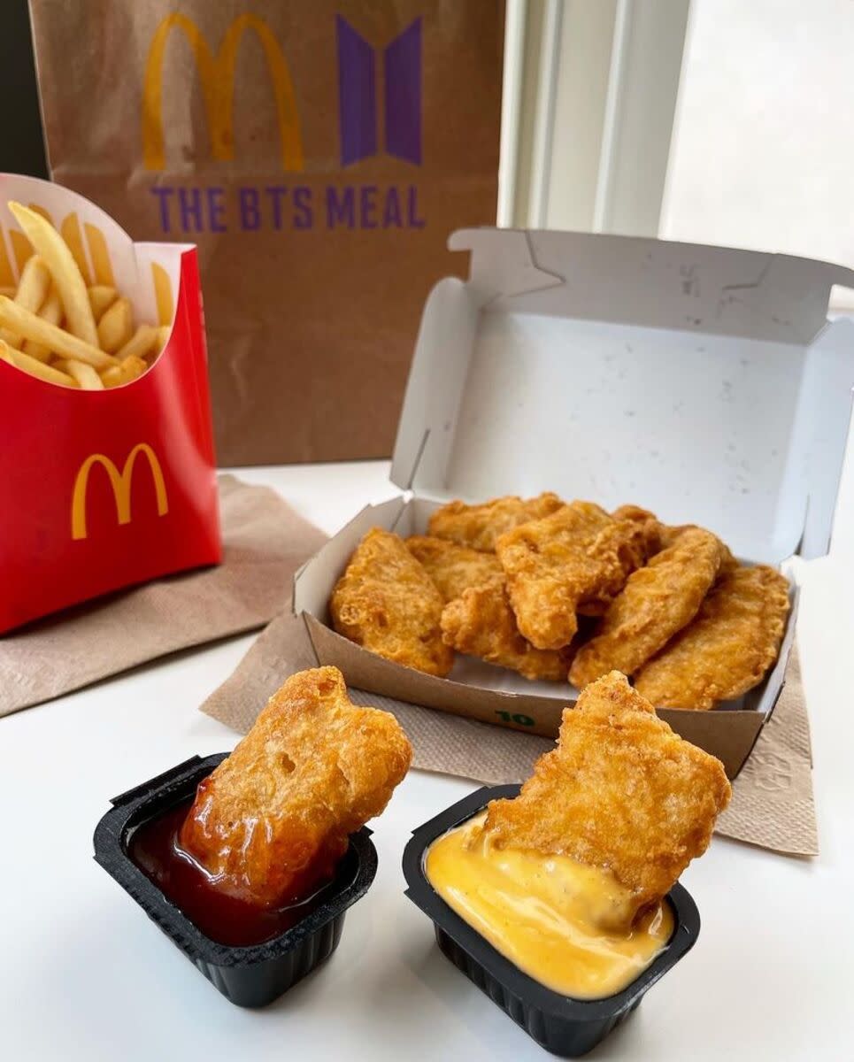 McDonald's BTS Meal, two chicken McNuggets in two different sauces with rest of chicken McNuggets on a napkin behind it, medium fries on a napkin with brown bag in the background, all on a white table