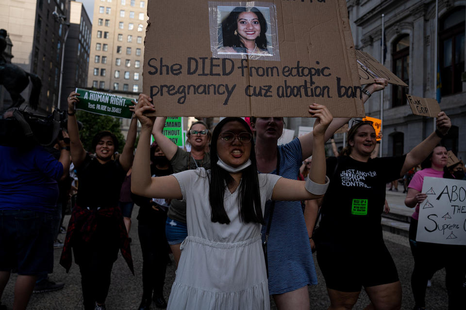 Protesters march in the street, with one holding up a sign with a photo of a woman and the text "She died from ectopic pregnancy cuz abortion ban"