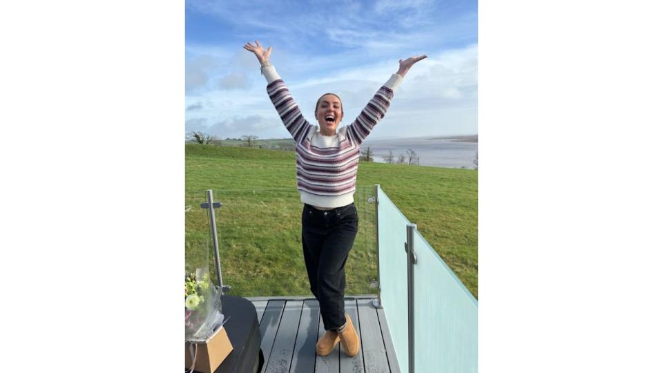 Amy Dowden on a balcony in front of grassy field with her hands in the air