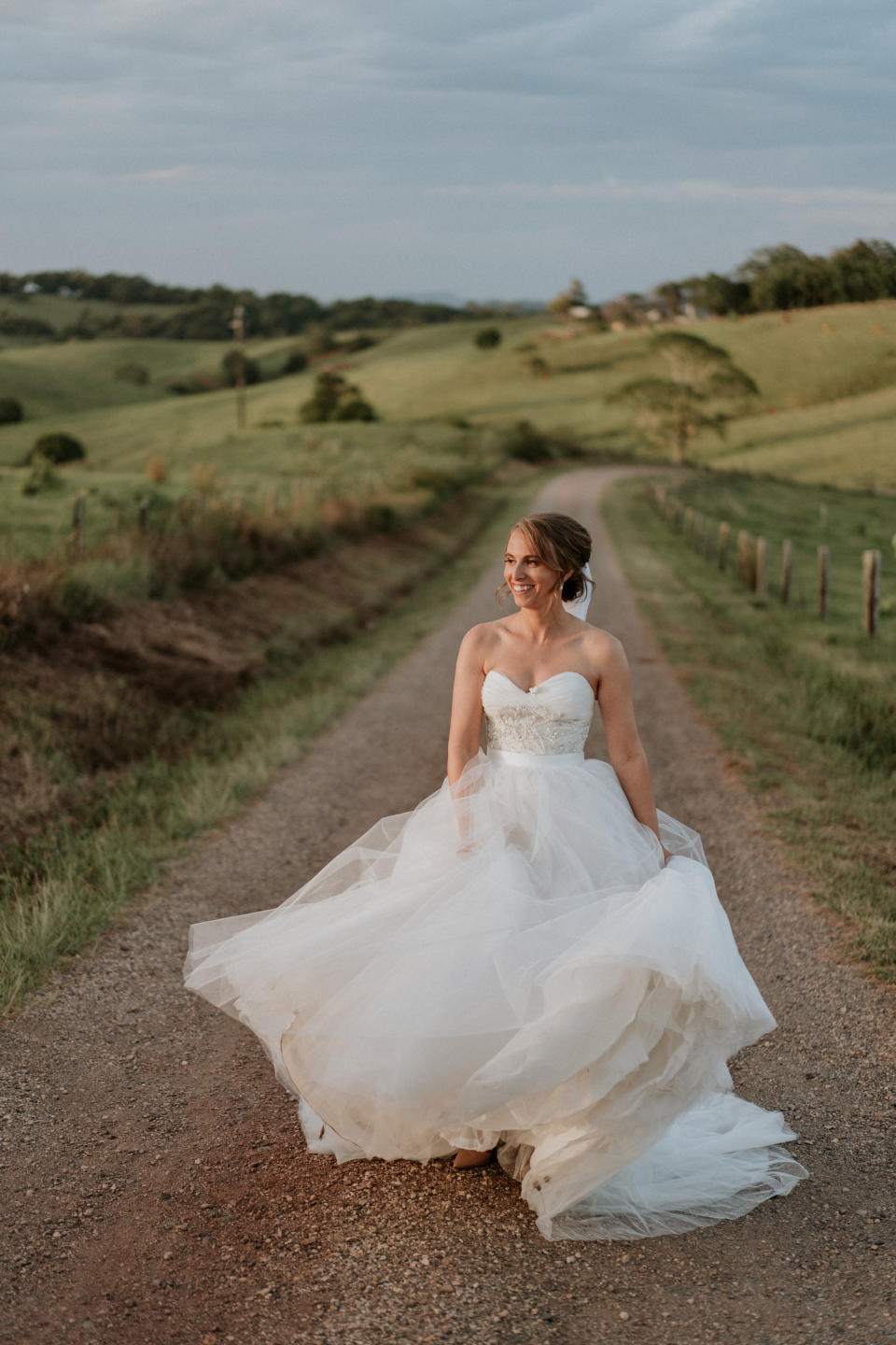 The bride, standing on a dirt road between green rolling hills, smiles as she plays with the skirt of her ball gown.&nbsp; (Photo: <a href="https://www.jamesday.com.au/" target="_blank">James Day Photography</a>)