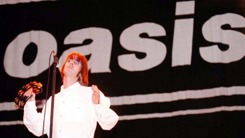 Liam Gallagher holding a tambouring on stage in front of a giant Oasis banner at Knebworth in 1996