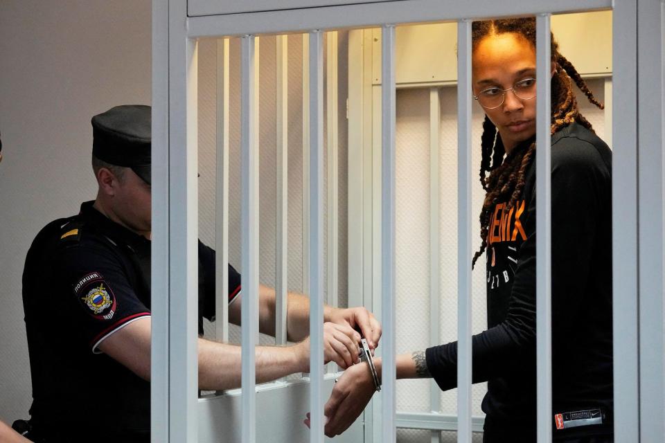 Brittney Griner stands in a cage as a Russian official adjusts her handcuffs.