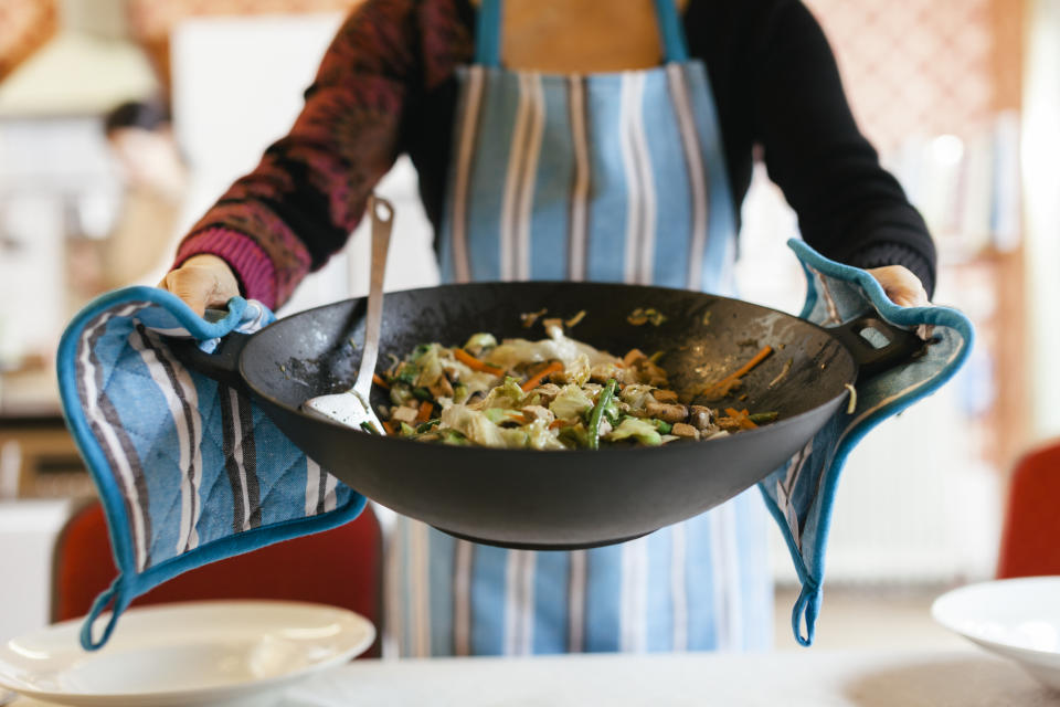 A cooking Wok  filled with a steamy dish is being held by someone wearing oven gloves. The dish consists of several different vegetables and tofu. The person holding the pan is wearing a kitchen apron.