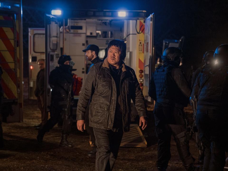 benedict wong as da shi in three body problem. he's a man in dark clothing, standing amid various armed personnel and several ambulances