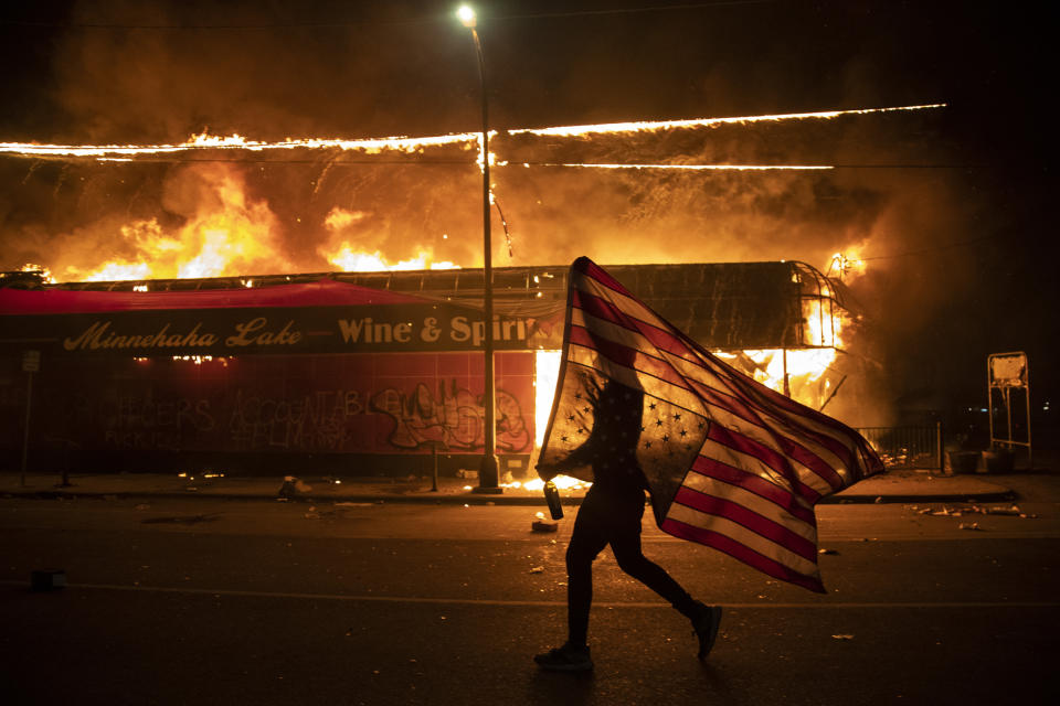 Un manifestante lleva una bandera estadounidense invertida frente a un edificio en llamas, el 28 de mayo de 2020 en Minneapolis. Forma parte de una serie de fotos de The Associated Press que ganó el Premio Pulitzer 2021 a la fotografía de noticias de último momento. (AP Foto/Julio Cortez)
