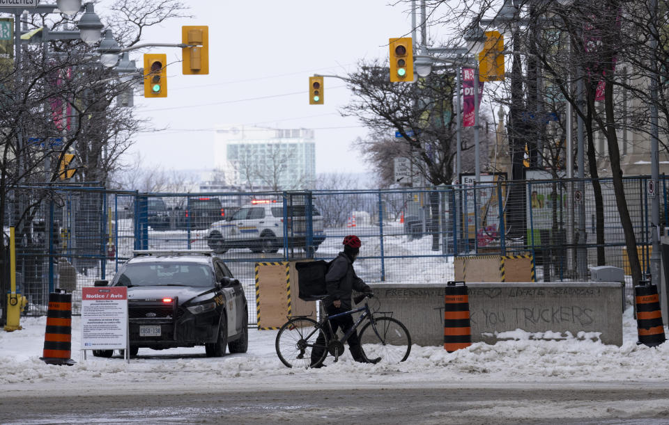A cyclist pushes his bike across an intersection past temporary fencing as police watch from their car, Wednesday, Feb. 23, 2022 in Ottawa. Ottawa protesters who vowed never to give up are largely gone, chased away by police in riot gear in what was the biggest police operation in the nation’s history. (Adrian Wyld /The Canadian Press via AP)