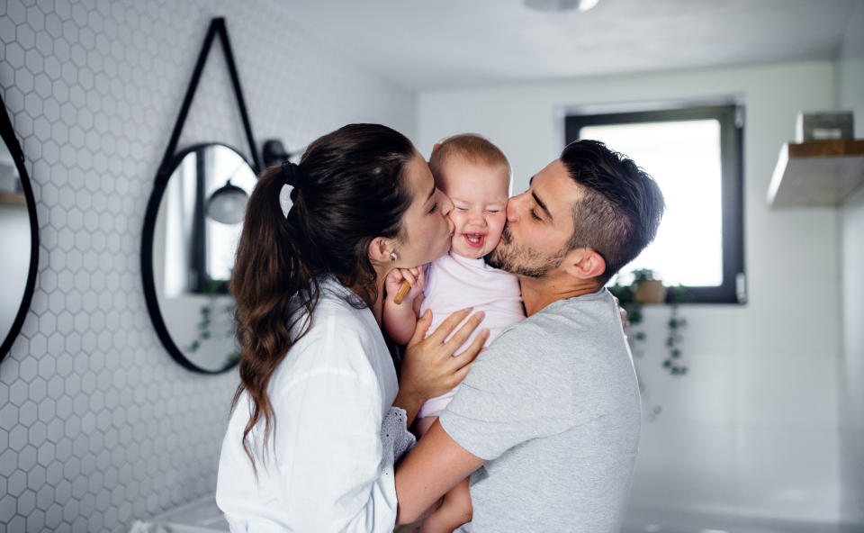 Two parents lovingly kiss a toddler's cheeks from either side in a home bathroom