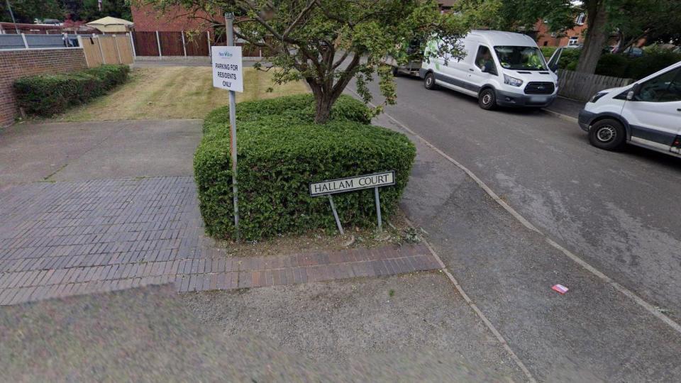 Streetview picture showing a Hallam Court road sign and two white vans parked on the road, in Ilkeston