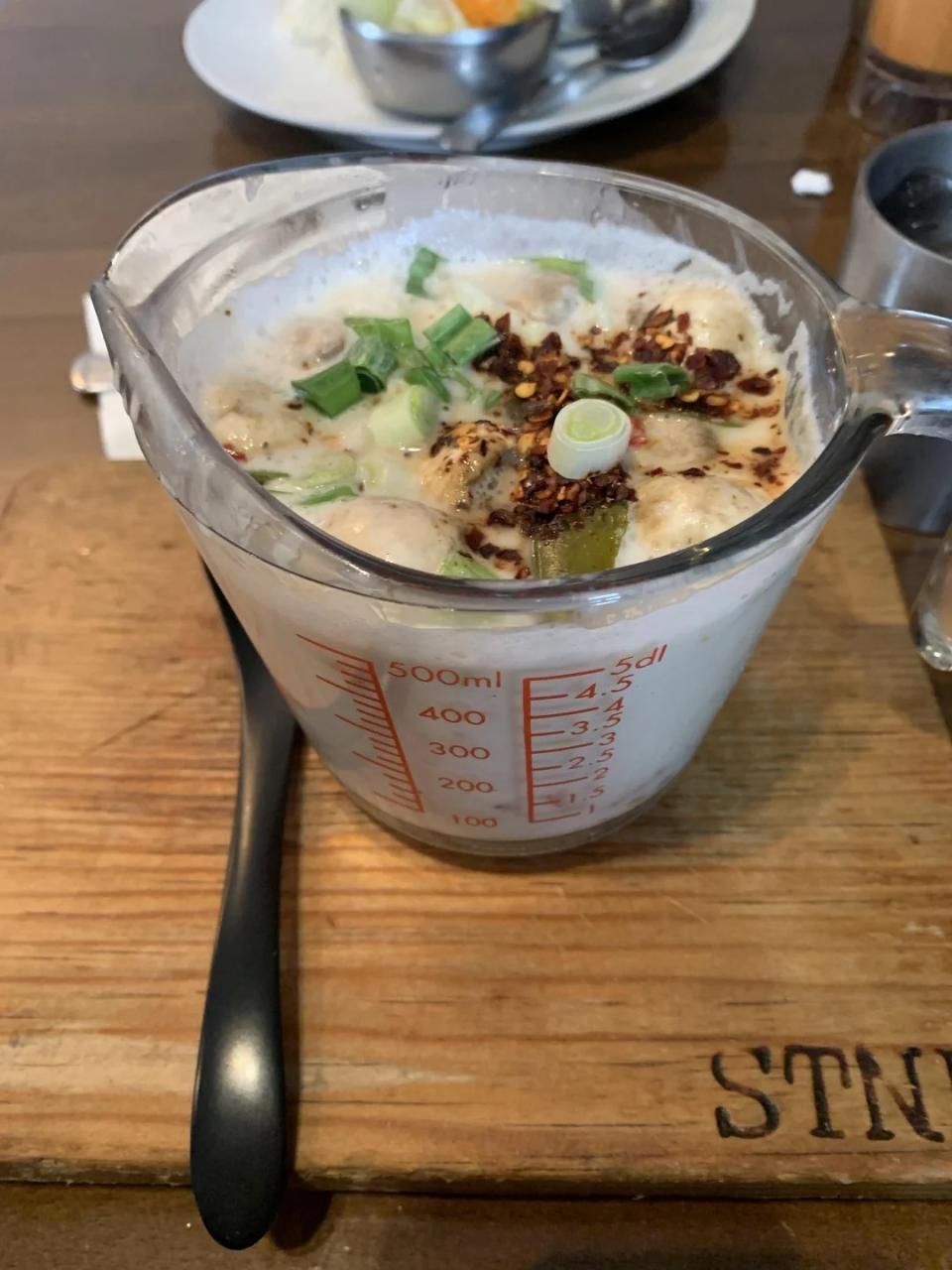 Asian-inspired soup with green onions, tofu, and chili flakes in a liquid measuring cup placed on a wooden board and spoon.