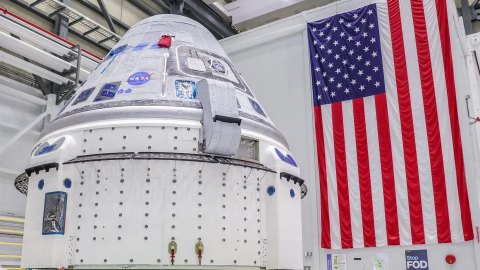 a grey and blue cone-shaped spacecraft sits with its small end up in a white hangar beneath a large American flag