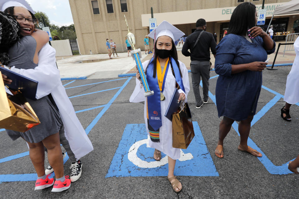 Amber Nguyen, valedictorian of the New Orleans Charter Science and Math High School class of 2020, wears a mask as she walks past a classmate hugging a family member, after receiving her diploma and valedictorian trophy, during a drive-in graduation ceremony as a result of the COVID-19 pandemic, outside Delgado Community College in New Orleans, Wednesday, May 27, 2020. Students and family got out of their cars to receive diplomas one by one, and then held a parade of cars through city streets. (AP Photo/Gerald Herbert)