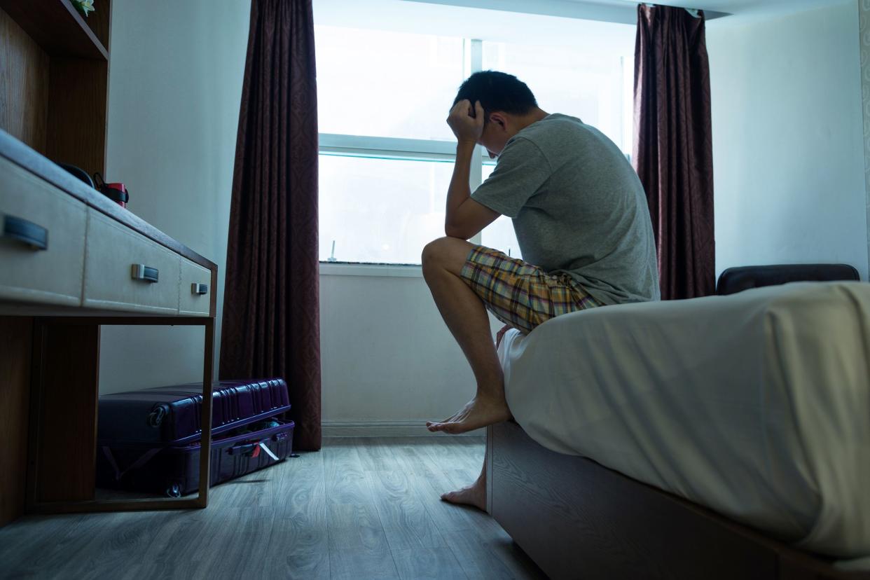 Distraught man sitting at edge of bed with his head in the his hands, bed on the right, desk on the left, in the foreground, a piece of carry-on luggage on the floor, a window with open curtains in the background, dark blue-toned image