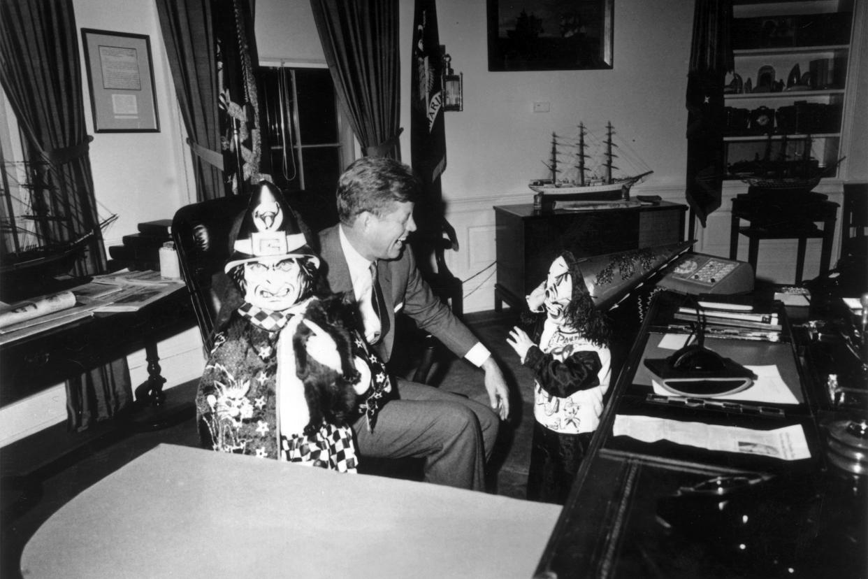 US President John F Kennedy (1917 -1963) sits behind his desk, laughing as his children Caroline and John Jr (1960 -1999), show him their Halloween costumes
