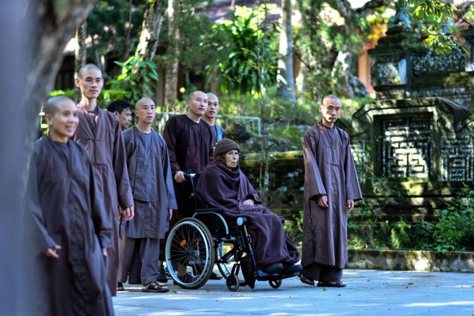 Buddhist monk Thich Nhat Hanh on a wheelchair wearing a purple robe and surrounded by monks in similar robes.