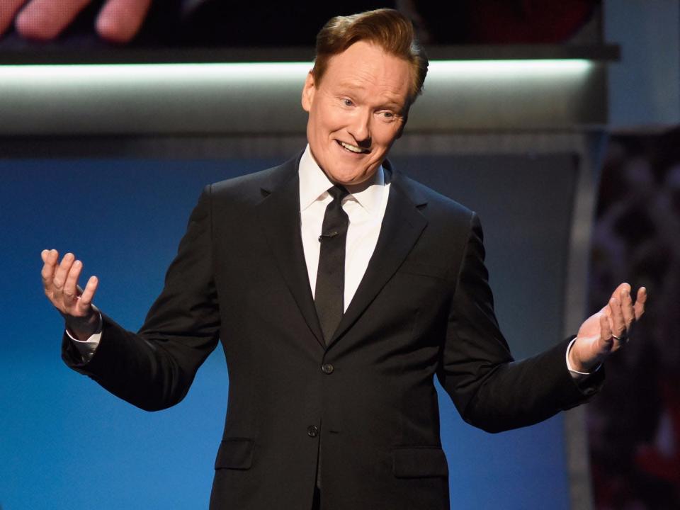 Conan O'Brien wears a black suit and gestures with a shrug