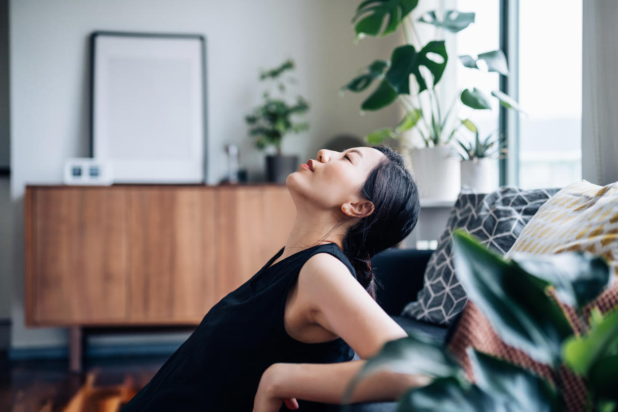 mindfulness Portrait of a beautiful young Asian woman relaxes by the sofa in the living room, with her eyes closed and head up, enjoying the tranquility during leisure time at cozy home