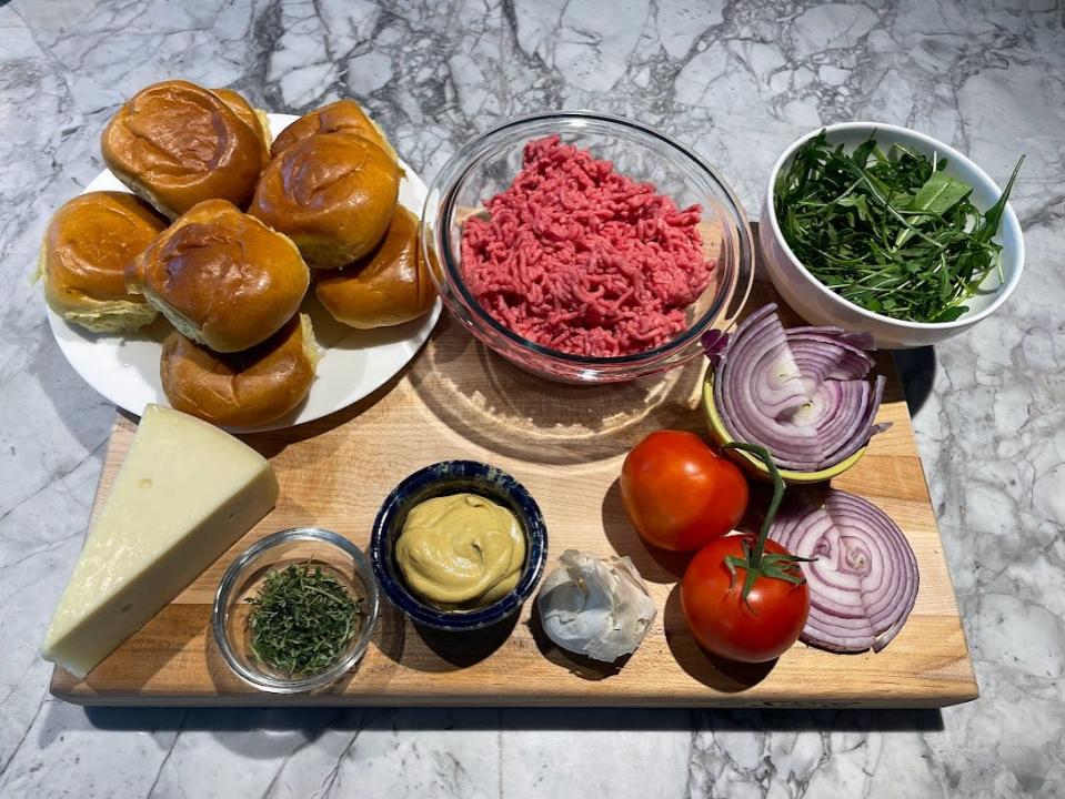 A cutting boards with hamburger buns, ground meat, arugula, mustard, garlic, tomatoes, onion slices, and a block of cheese laid out