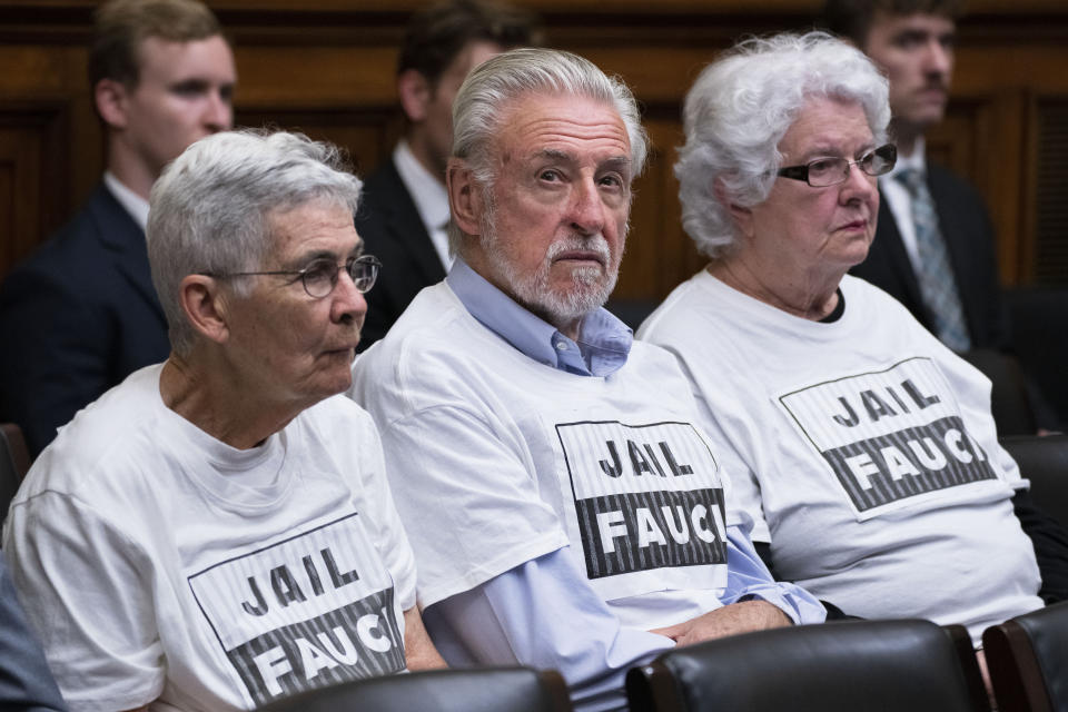 A gray-haired man flanked by two gray-haired women watch the hearing, all wearing white T-shirts saying: Jail Fauci..