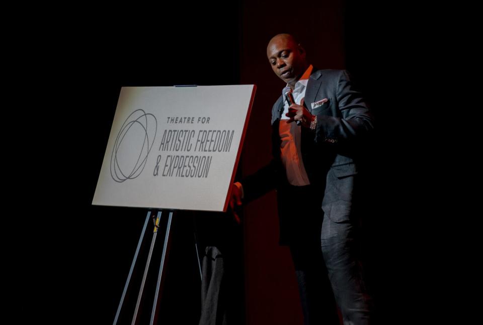 A man stands onstage next to a sign that says Theatre for Artistic Freedom and Expression