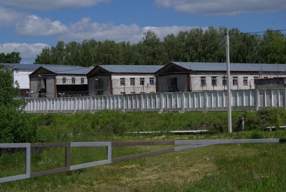 Three uniform cell blocks behind a barrier mounted with a wire fence, in a wooded setting.