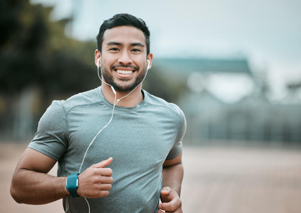 A man jogging outdoors, smiling at the camera, wearing a t-shirt, earphones, and a fitness tracker